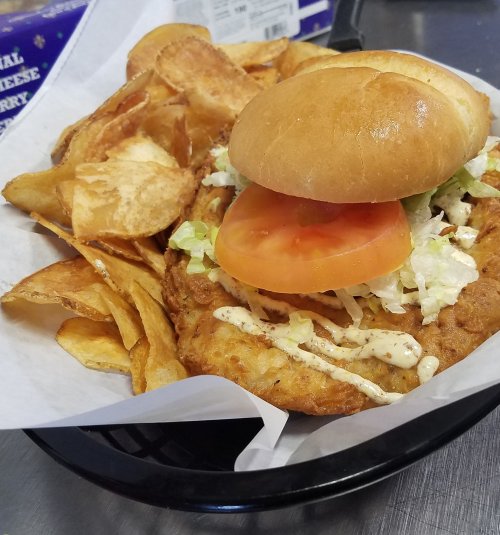 A crispy chicken sandwich with lettuce and tomato, served with a side of homemade potato chips on a black plate.