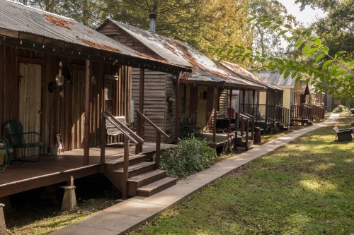A row of rustic wooden cabins with porches, surrounded by greenery and a stone pathway.
