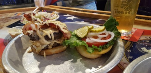 A burger with bacon, cheese, and toppings on a metal plate, alongside a fresh bun with lettuce, tomato, and pickles.
