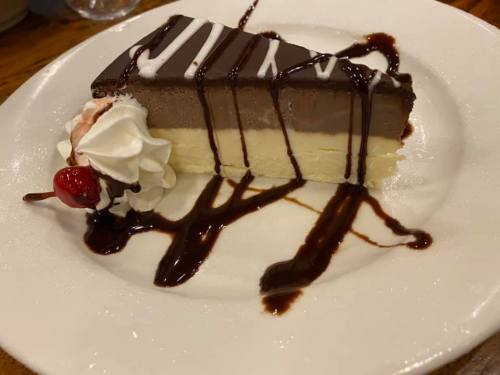 A slice of layered chocolate mousse cake topped with whipped cream, a cherry, and chocolate drizzle on a white plate.