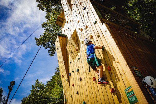 A child climbs a wooden rock wall at an outdoor climbing facility under a blue sky.