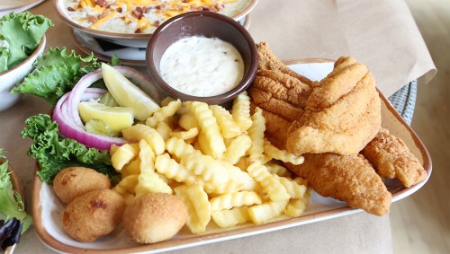A plate of fried catfish fillets, crinkle-cut fries, hushpuppies, and a side of tartar sauce with lemon and onions.