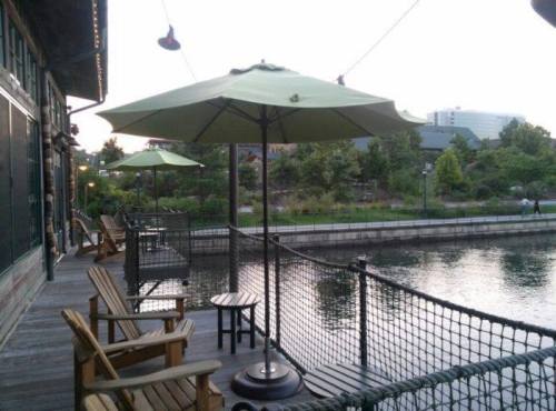A peaceful riverside patio with wooden chairs, green umbrellas, and a calm water view at dusk.