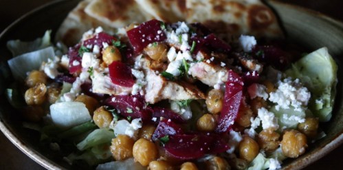 A bowl of salad with lettuce, chickpeas, beets, grilled chicken, feta cheese, and flatbread on the side.