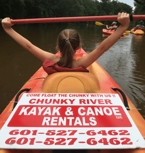 A girl in a kayak raises her arms, with a sign for Chunky River Kayak & Canoe Rentals visible behind her.