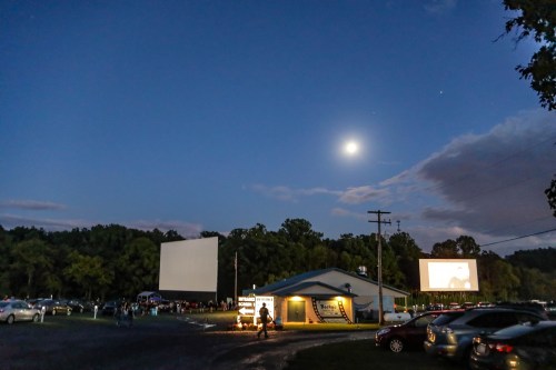 A drive-in movie theater at dusk, with a bright moon and two screens displaying films, surrounded by parked cars.