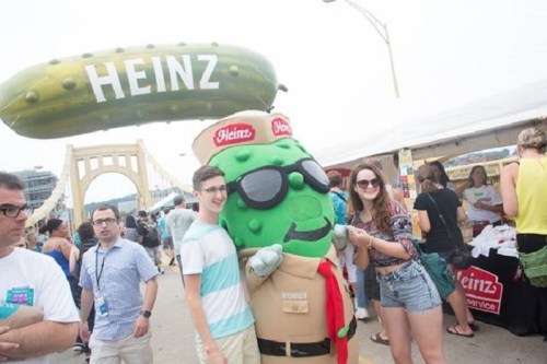 A giant pickle mascot poses with two smiling people at a festival, with a bridge and vendor tents in the background.