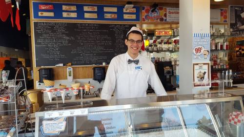 A smiling ice cream shop employee in a white uniform and bow tie stands behind the counter with ice cream displays.