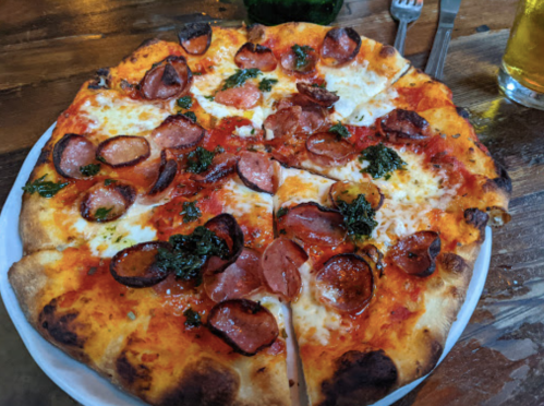 A freshly baked pizza topped with sliced sausage, cheese, and herbs on a wooden table.