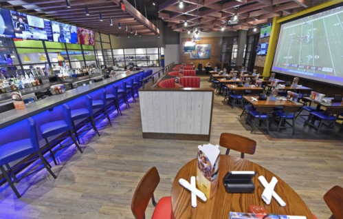 A modern restaurant interior with a bar, dining tables, and a large screen showing a sports game.