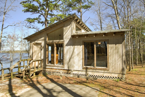A wooden cabin by a lake, surrounded by trees, with large windows and a small deck.