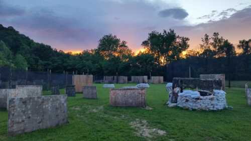 A paintball field with wooden barriers and a stone structure, set against a colorful sunset and green trees.