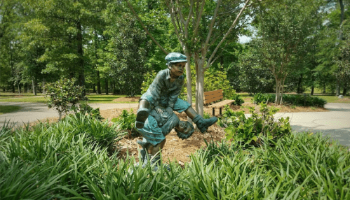 A bronze statue of a boy in a cap, playfully crouching among greenery in a park setting.