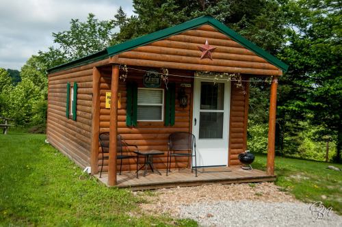 A small log cabin with green shutters, a star decoration, and a porch with two chairs, surrounded by greenery.