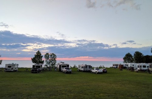 A serene campsite with RVs parked on green grass, overlooking a calm lake at sunset with colorful clouds.