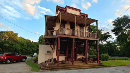 A three-story building with a porch, surrounded by greenery and parked cars, under a blue sky with clouds.