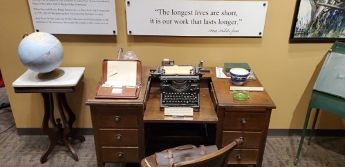A vintage typewriter on a wooden desk with a globe and books, accompanied by a quote on the wall about lasting work.