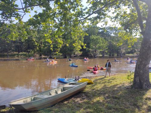 A serene river scene with kayakers paddling, a small boat on the shore, and lush greenery surrounding the water.