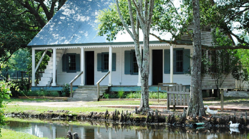 A charming white house with a sloped roof, surrounded by trees and a small pond, featuring steps and open doors.