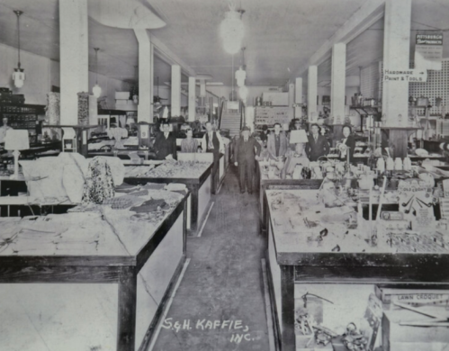 Historic interior of a general store, featuring merchandise displays and customers in early 20th-century attire.