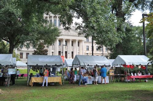 A lively outdoor market with tents, people browsing, and a historic building in the background surrounded by trees.