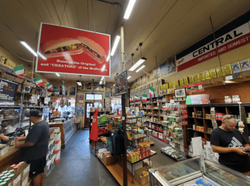 Interior of a vibrant grocery store with shelves stocked with various products, featuring a large sign about "CREATORS of the Muffuletta."