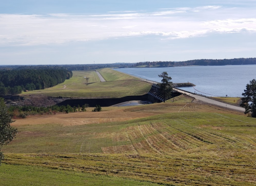 A wide view of a dam with a road on top, surrounded by grassy fields and a calm lake under a clear sky.