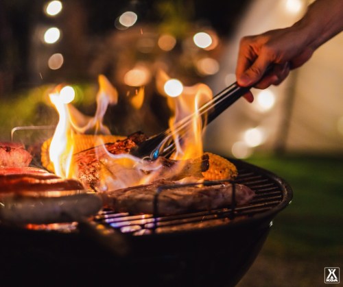 A hand using tongs to grill meat over an open flame, with a blurred background of lights and greenery.