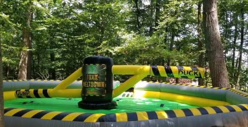 A colorful inflatable game called "Toxic Meltdown" set up in a wooded area, featuring a green pool and safety barriers.