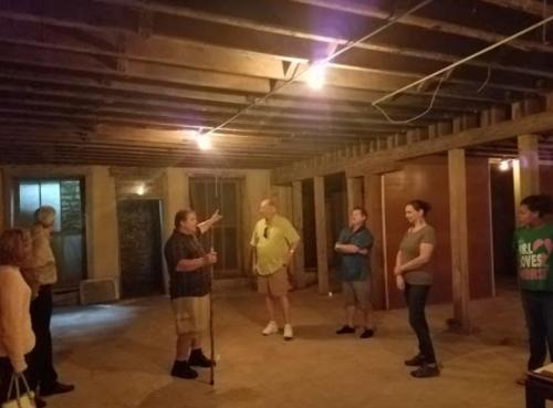 A group of people stands in a dimly lit basement, listening to a speaker with a cane. Exposed beams and walls are visible.