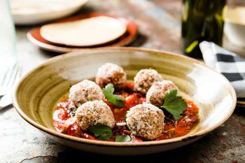 A bowl of meatballs in tomato sauce, garnished with herbs, served on a rustic table with plates in the background.