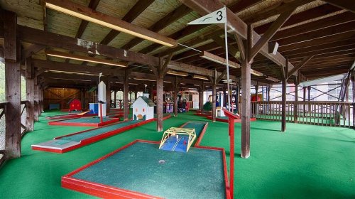 Indoor mini-golf course with colorful obstacles and a green carpet under a wooden ceiling.