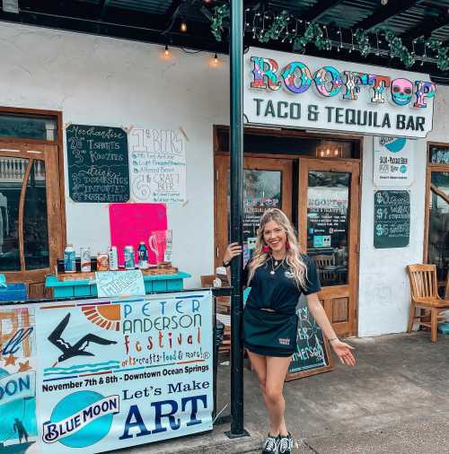 A smiling woman stands outside a taco and tequila bar, showcasing festival signs and a vibrant storefront.