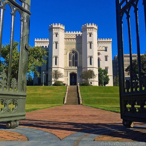 Historic castle-like building viewed through ornate gates, surrounded by green lawns and a clear blue sky.