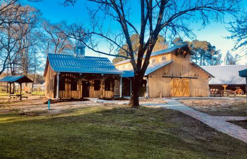 A rustic wooden barn and pavilion surrounded by trees and a clear blue sky, with a pathway leading through the grass.