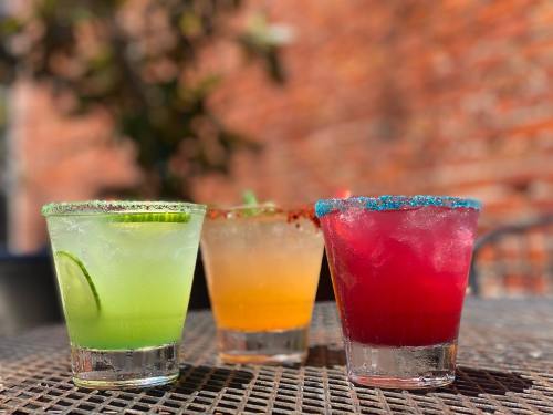 Three colorful cocktails with garnishes on a textured table, set against a blurred brick background.