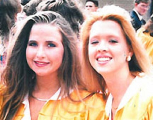 Two young women in yellow graduation gowns smile at the camera, surrounded by a crowd in the background.