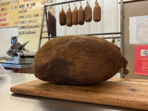 A large, brown, burlap-wrapped item sits on a wooden cutting board in a deli with hanging meats in the background.