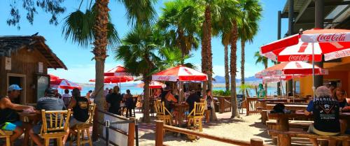 A lively beachside scene with palm trees, red umbrellas, and people enjoying food and drinks at outdoor tables.