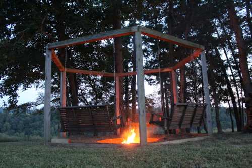 A wooden swing structure surrounds a fire pit, set in a grassy area with trees in the background at dusk.