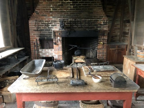 A rustic kitchen with a brick fireplace, wooden table, and various cooking tools and utensils scattered around.