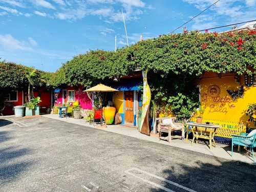 Colorful buildings with vibrant plants, outdoor seating, and a sunny sky in a charming courtyard setting.