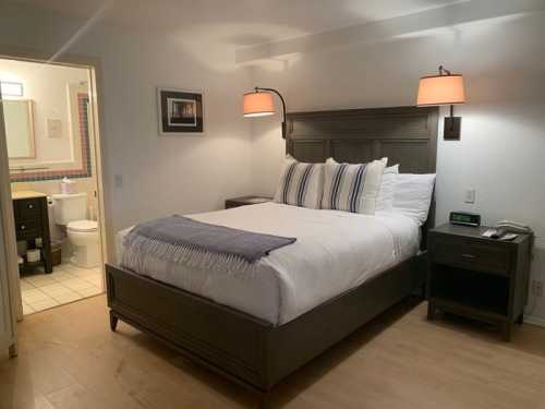 Cozy hotel room featuring a queen bed, bedside lamps, and an adjoining bathroom. Light wood flooring and modern decor.