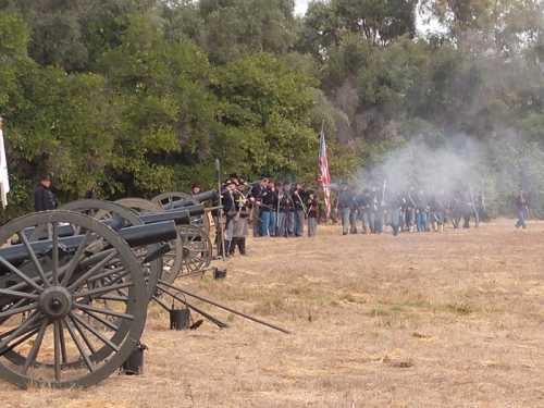 Civil War reenactors with cannons and an American flag, set in a grassy field with smoke in the background.