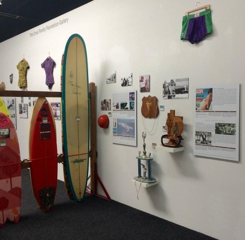 A gallery wall featuring surfboards, vintage swimwear, and historical photos related to surfing culture.