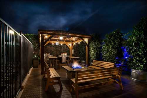 A cozy outdoor seating area with a fire pit, surrounded by greenery, illuminated by soft lights at night.