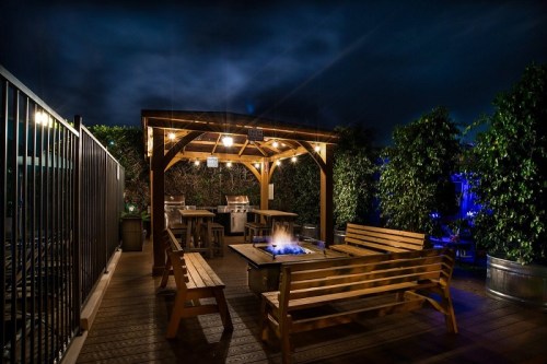 A cozy outdoor seating area with a fire pit, surrounded by greenery, illuminated by soft lights at night.