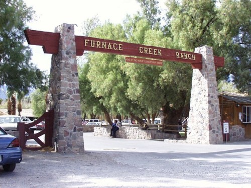 Entrance to Furnace Creek Ranch, featuring a stone archway and wooden sign, surrounded by trees and parked cars.