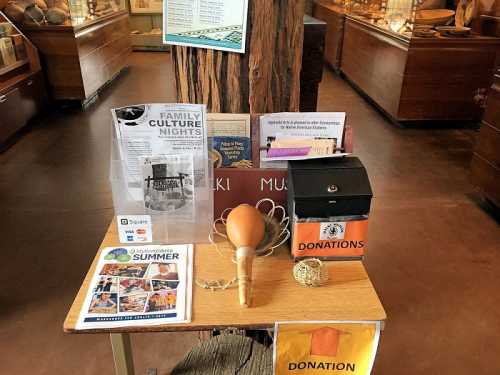 Display table in a museum featuring donation box, flyers for events, and cultural materials.