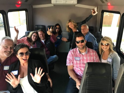 A group of people smiling and posing inside a bus, some waving and showing excitement. Name tags visible on their shirts.